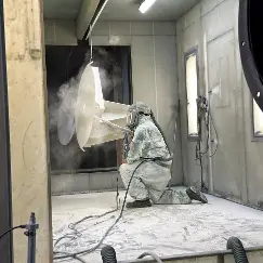 Worker in full protective suit and respirator spraying material inside a paint/spray booth, with plastic sheeting and machinery around.