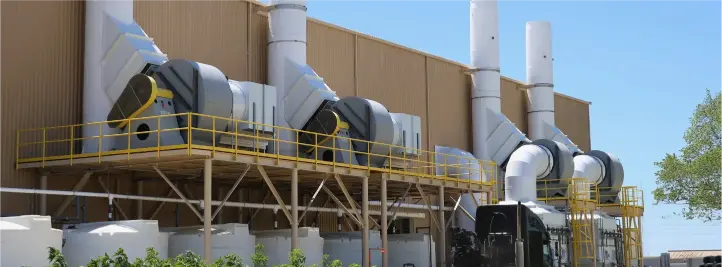 Industrial exhaust ventilation system on an exterior building with large ducts, circular vents, and yellow safety railings along a raised catwalk under a clear blue sky