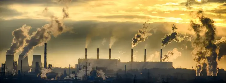 Industrial plant with tall smokestacks releasing dark smoke at sunset.