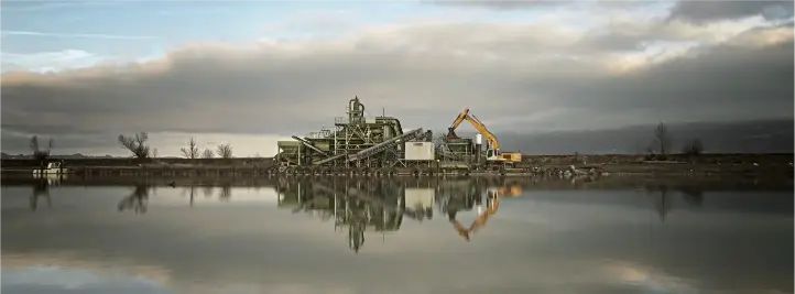 Industrial processing facility on the shore with a yellow excavator, reflected in a calm body of water under cloudy sky