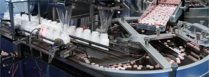 Automated bottling line with white bottles on a conveyor and pink and white tablets moving through filling equipment.
