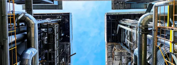 View upward between two tall industrial buildings with large metal pipes and catwalks, blue sky overhead.