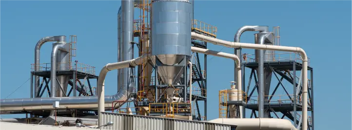 Industrial refinery with a large blue vertical vessel and a network of silver pipes against a clear sky, with yellow railings visible.