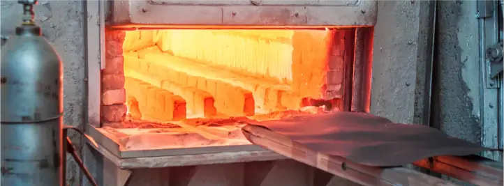 Industrial forge with a glowing orange furnace interior and metal bars on a tray entering the heat chamber near a propane tank on the side.