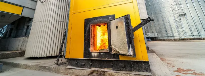 Open industrial furnace door with bright flames visible inside a yellow housing, outdoors beside a modern building.