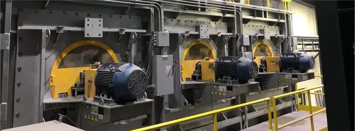 Row of industrial machines with yellow circular doors and blue motors in a factory, with a safety railing in front.