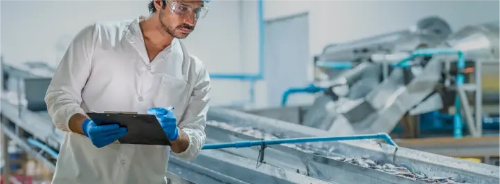 Lab technician in safety goggles and blue gloves inspects a stainless steel conveyor, holding a clipboard in a bright industrial setting.