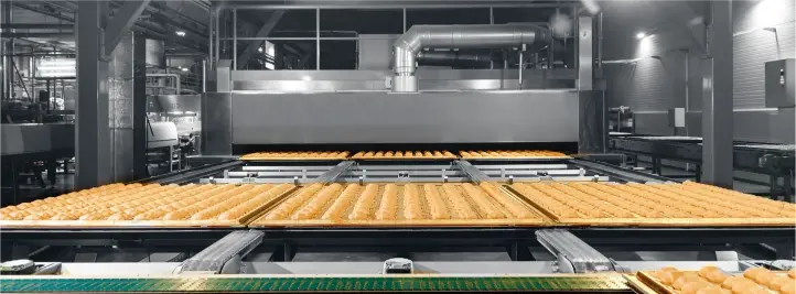 Industrial bakery production line with trays of golden pastries moving along a conveyor toward a large oven.