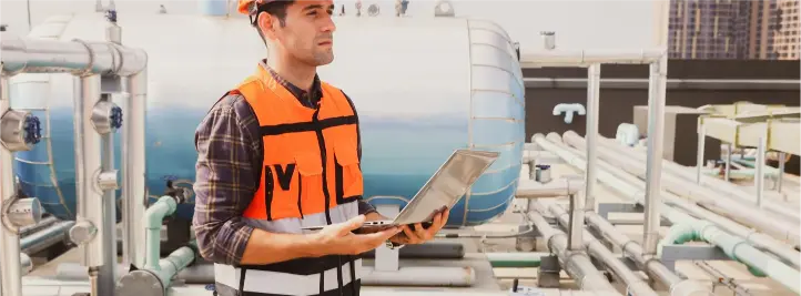 Male technician in orange safety vest and helmet stands among industrial pipes, holding a tablet on a rooftop inspection site.