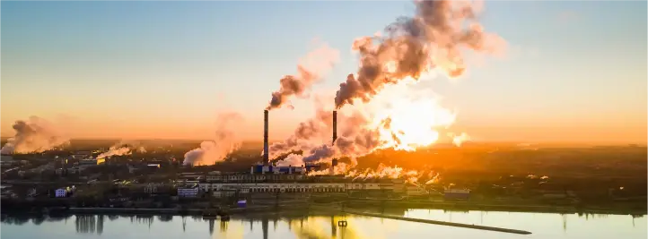 Industrial power plant with tall smokestacks emitting smoke at sunset over a river, reflections on the water.