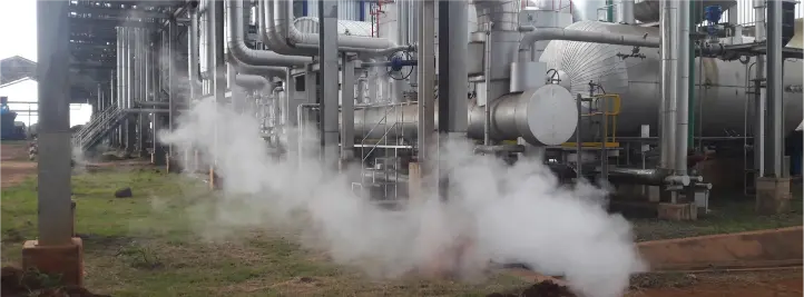 Industrial plant with piping and steam venting from ground near equipment pieces behind it in a refinery-like setting, daytime outdoor scene.