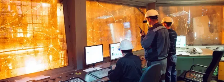 Three technicians in hard hats monitor computers at a control room with orange safety glass walls.