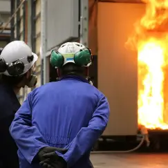 Two workers in hard hats and blue coveralls watch a blazing fire inside a metal chamber after a safety alert/inspection