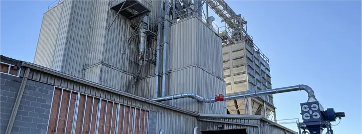 Industrial plant exterior with tall corrugated metal silos, piping, and a red valve in the foreground under a clear blue sky.