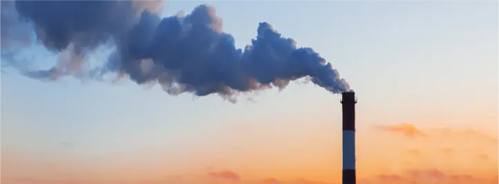Industrial smokestack emits a thick plume of dark smoke against a orange-pink sunset sky at dusk.