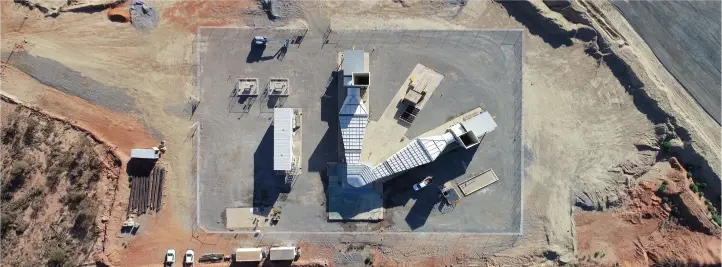 Aerial view of an industrial site on a concrete pad, featuring a tall central tower and several connected rectangular structures, with dirt surroundings.