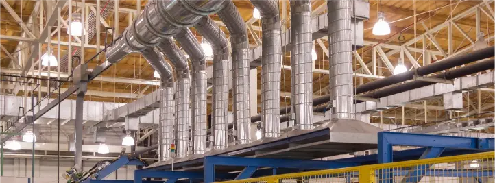 Interior of a factory ceiling with a network of shiny metal ductwork and pipes above blue machinery and yellow safety rails