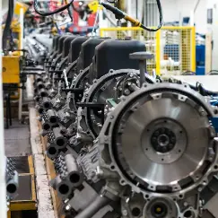 Row of large metal engines mounted on a manufacturing line, with yellow safety rails and equipment in the background.