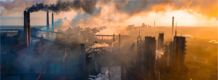 Industrial complex with tall smokestacks emitting dark smoke at sunset, haze filling the air.