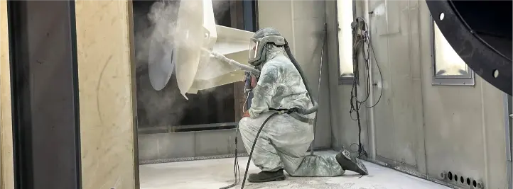 Hazmat-suited worker kneeling and using a spray gun to apply coating inside an industrial spray booth.