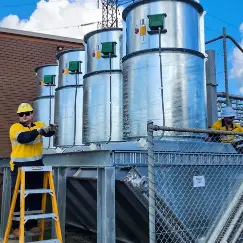 Three large stainless steel industrial tanks on a raised platform, with two workers in safety vests and hard hats inspecting equipment behind a chain-link fence.