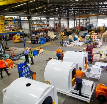 Factory floor with workers in orange vests assembling large white cylindrical metal housings on a production line.