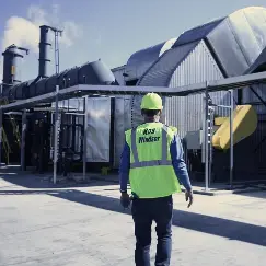 Worker in a high-visibility vest and hard hat walks toward large industrial equipment behind a fence on a plant site.