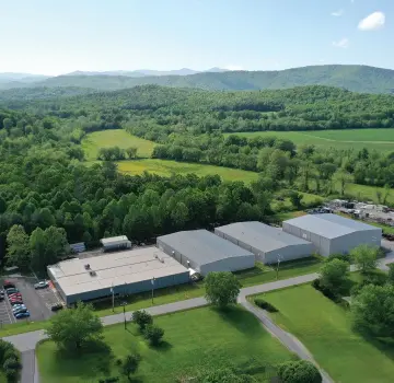 Aerial view of a business park with gray warehouses, a parking lot, and surrounding green fields and trees. in distance hills.