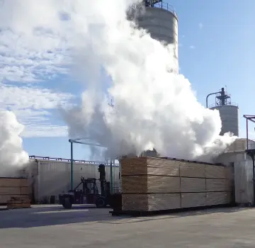 Forklift handling stacked lumber at an industrial yard, with large boards and white exhaust or steam rising in the background.