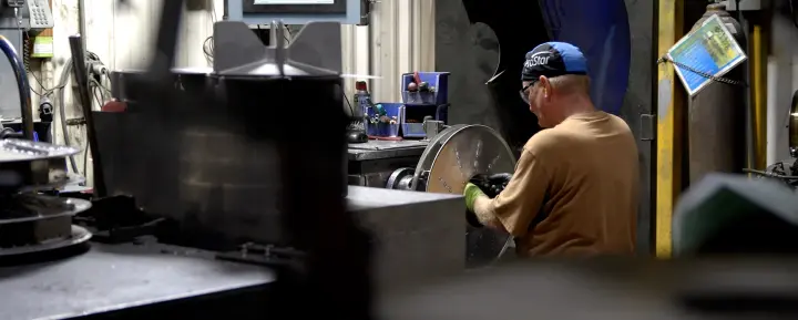 Worker wearing a blue cap and green gloves operates a large metal wheel on a machine in a busy manufacturing workshop.