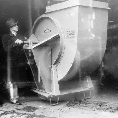 Man standing beside a large industrial rotary blower with a circular intake and an open front door in a workshop.