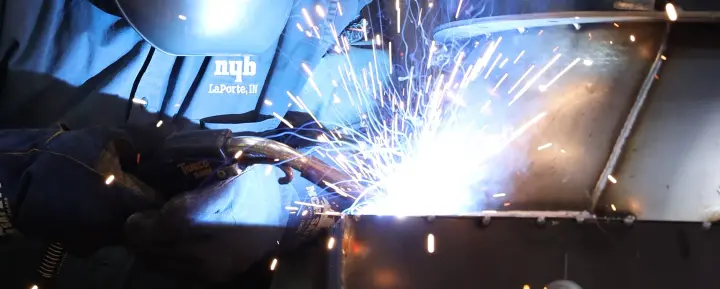Welding metal with bright sparks; worker in blue protective jacket and helmet uses a welding torch.