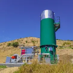 Green vertical industrial storage tank with a metal staircase and platform, set on a sandy hill under a clear blue sky, with auxiliary equipment nearby.