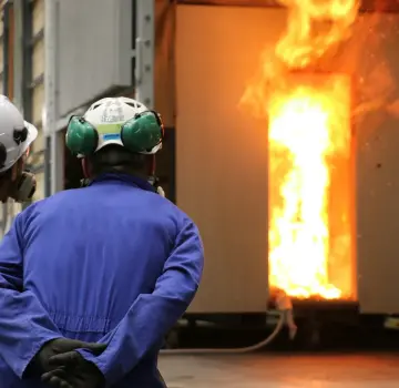 Two workers in blue coveralls and hard hats stand with their backs to the camera, watching a blazing furnace. Their PPE includes ear protection and gloves.