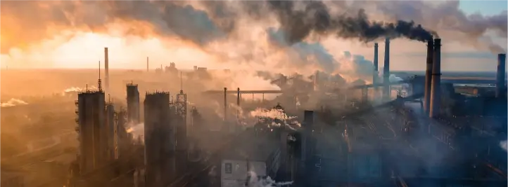 Industrial facility with tall smokestacks emitting smoke, creating a hazy skyline at sunset.