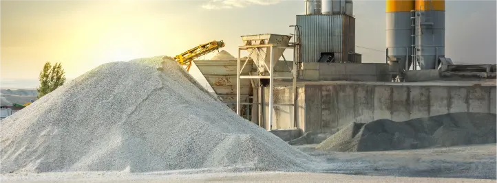 Industrial quarry or cement plant scene with large piles of gravel, conveyors, and silos at sunset.
