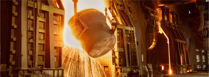 Molten metal being poured from a crucible into a mold in a steel foundry with a bright orange glow and sparks.