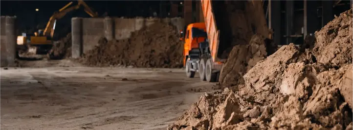 Night construction site with dirt piles and an orange dump truck under dim lighting at night in the foreground.