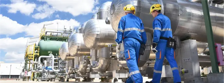 Two workers in blue coveralls and yellow hard hats inspect large silver industrial pipes outdoors under a blue sky.