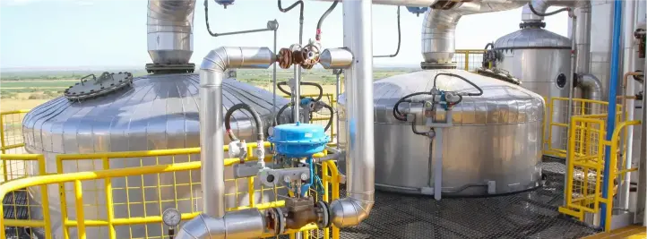 Outdoor industrial processing facility with large stainless steel reaction tanks, network of silver pipes, and yellow safety railings across a metal walkway.