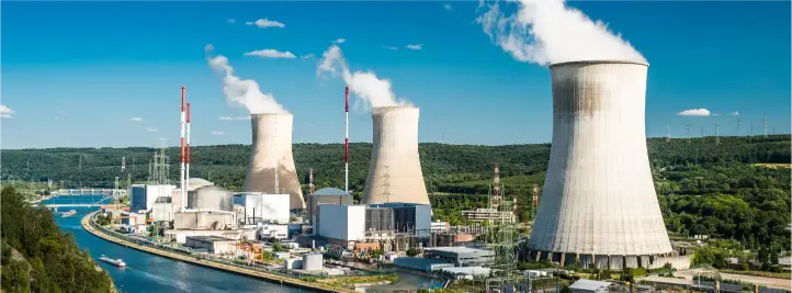 Industrial power plant with three cooling towers emitting steam beside a waterway and greenery