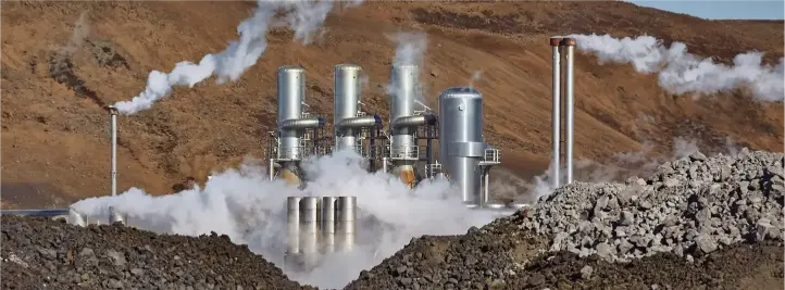 Industrial facility with multiple smokestacks releasing steam in a barren quarry setting, brown hills in the background.