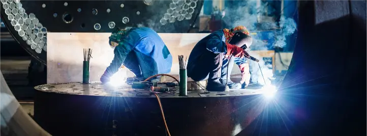 Two welders in PPE bend over a large metal plate, sparks flying as they weld in a dim workshop.