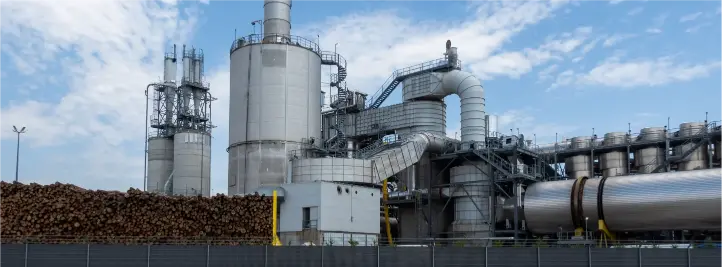 Industrial chemical plant with tall silo towers, connected pipes, and a log pile in the foreground.