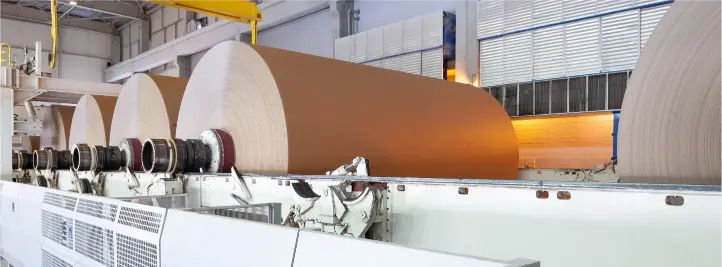Large rolls of brown paper on a conveyor in a paper mill, with industrial machinery and overhead cranes nearby