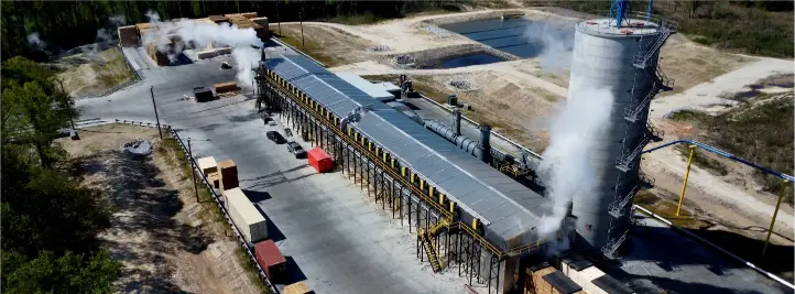 Aerial view of a large industrial processing plant: a long gray structure with yellow railings and steam plumes rising from vents at either end.