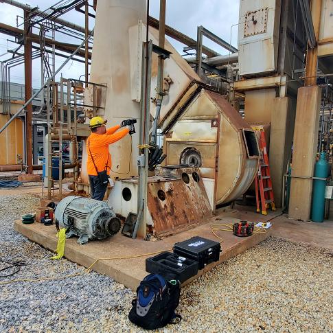 Worker in orange safety gear and a yellow hard hat operates a large, rusted industrial machine outdoors, with tools and cables nearby.