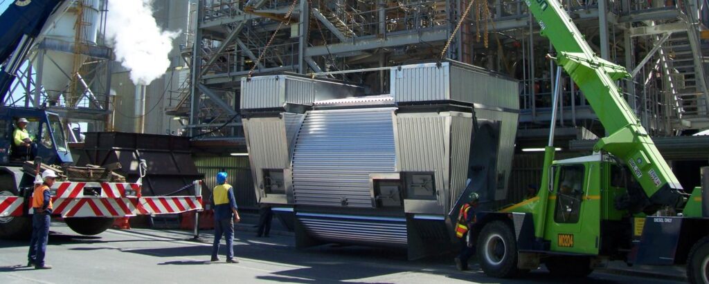 Industrial construction site with large silver machinery being moved by a green crane and workers guiding the load.