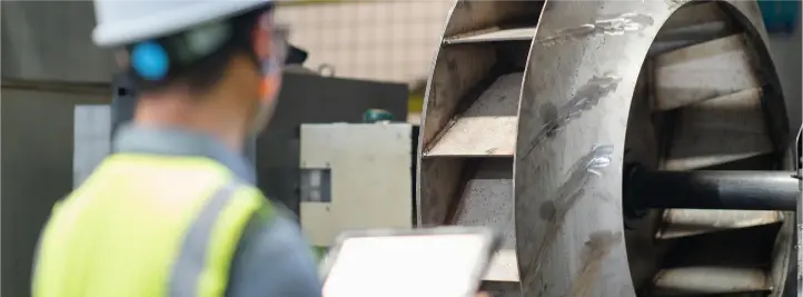 Worker in a hard hat and high‑visibility vest uses a tablet to inspect a large industrial turbine rotor.