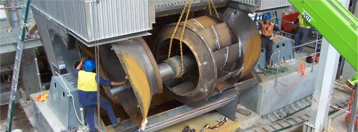 Large industrial rotor or turbine drum suspended by a crane, with workers in safety gear nearby.